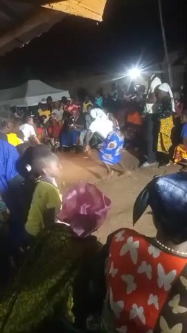 The video shows a nighttime gathering of people outdoors, likely in a village setting. A group of individuals is centered in a clearing, with a crowd of spectators surrounding them. The central figures appear to be engaged in some form of performance or ritualistic activity, possibly a dance or a ceremonial struggle. One person, wearing a white top and a blue and white patterned skirt, is in motion, with another figure in a white outfit close by. The ground is dusty, suggesting it is a natural, unpaved area. Tents or canopies are visible in the background, along with artificial lighting that illuminates the scene. The spectators are observing the central action with attention. The overall atmosphere appears lively and communal.
