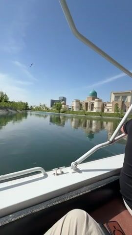The video captures a serene daytime scene from the perspective of a passenger on a small white boat, slowly navigating a calm body of water, likely a canal or lake, under a clear blue sky with sparse clouds.

In the foreground, parts of the boat's white frame, railings, and the lower body of a passenger (wearing light-colored pants) are visible. The main subject is a man with short, graying hair, viewed from behind. He is wearing a black short-sleeved polo shirt with red and white trim on the collar and sleeves, and light-colored pants. He is seated at the boat's helm, actively piloting the vessel, with an orange life vest visible on the deck near his steering console. The gentle hum of the boat's motor can be heard throughout the clip.

The background showcases a landscaped shoreline. A prominent light-colored building with a large, distinctive light green or turquoise dome and arched windows stands directly across the water, its reflection clearly visible on the calm surface. Adjacent to this building are other modern-looking structures. As the boat proceeds and the camera pans slightly, more distant contemporary buildings and lush green trees lining the banks come into view, suggesting a developed and well-maintained area. The overall atmosphere is peaceful and leisurely, characteristic of a scenic boat tour in a park or waterfront district, consistent with Kuchtumgali, Uzbekistan.