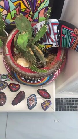 The video shows a close-up of a collection of painted rocks displayed on a white surface next to a potted cactus in a decorated planter. The rocks are various shapes and sizes, each with a unique hand-painted design. Some feature abstract patterns, while others depict stylized faces or symbolic imagery. The planter itself is elaborately decorated with colorful paint and possibly beads or studs. In the background, a vibrant, abstract artwork is visible. To the right, a glass surface reflects a blurry image of objects, including a Heineken bottle opener and what appears to be a small, round container with colorful decorations. The lighting suggests an indoor setting, possibly a craft area or a display space.