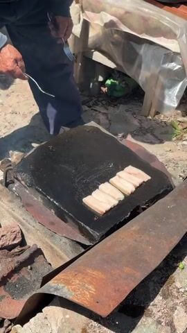A person, wearing a blue shirt and dark pants, is cooking pieces of pork belly on a large, flat black griddle. The griddle is placed over a makeshift outdoor stove, constructed from metal and bricks, with smoke rising from beneath it. The person is using a spatula and fork to turn the meat. In the background, a table is covered with a plastic sheet, and a plate with more raw meat is visible on the table. The scene appears to be outdoors, with dirt and scattered debris on the ground.