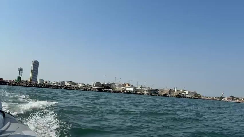 A wide shot shows a boat moving through blue water, creating a wake. In the background, a coastline is visible under a clear blue sky. The coastline features a variety of buildings, including a tall, slender skyscraper on the left, several lower structures, and what appear to be port facilities with cranes and scaffolding. A rocky breakwater separates the sea from the land. The overall impression is of a coastal city or town seen from the water.