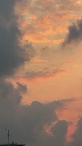 The video shows a time-lapse of a cloudy sky at sunset. The clouds are a mix of dark gray and light orange and pink hues. In the lower left corner, the top of a white building with a few windows is visible, along with an antenna. In the distance, a dark mountain range can be seen. The overall impression is of a dramatic and beautiful sunset.