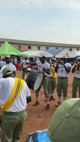 A video captures an outdoor scene of a marching band practicing. The band members are dressed in green trousers and white t-shirts with yellow and green sashes. Some members are playing drums, including a large bass drum. Others are playing brass instruments such as trumpets. The leader of the band, in the center, is holding a drumstick and a mace, both of which are decorated with a silver sphere at the top. The background shows tents and buildings, suggesting an organized event or gathering. The ground is covered in dirt. The camera is positioned at a low angle, looking up at the band members. The video is short, lasting only a few seconds, and features the sounds of the marching band.
