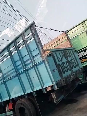 The video displays a close-up, shaky view of two trucks on a road. The primary truck in the foreground is a large, light blue flatbed truck with a tall, cage-like structure on its bed. The truck is angled, and its tires and part of its undercarriage are visible. In the background, a yellow truck is partially visible, appearing to be a box truck. The camera seems to be moving, possibly inside another vehicle, as evidenced by the reflections on the surface, which include what appears to be a dashboard and possibly other vehicles. The lighting suggests it is daytime. The scene appears to be in a congested area, as the trucks are close together, implying traffic or parking.