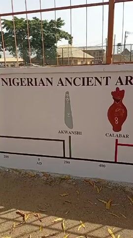 The camera pans across a wall displaying “NIGERIAN ANCIENT ART TRADITIONS”. The wall features a timeline beginning at 3000 BC and ending at 1400 AD. Images of Nigerian artifacts are displayed along the timeline, with labels including NOK, AKWANSHI, CALABAR, IGBO-UKWU, IFE, OWO, and TADA. The wall is painted white with black lettering and images. A fence runs along the top of the wall. A tree is visible behind the wall, as well as buildings and a vehicle. The ground in front of the wall is covered in gravel and leaves.