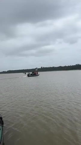 A wide shot captures a river under a cloudy sky. In the distance, a small boat carrying several people and two flags, one a blue and white flag with a sun and the other a green and red flag, navigates the water. The boat is moving from left to right across the frame. The banks of the river are lined with trees. The water is calm with small ripples. The camera perspective suggests the viewer is on another boat.