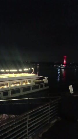 The video shows a nighttime view of a body of water with various lights reflecting on its surface. In the foreground, there is a section of a walkway or pier, constructed from metal.  A small boat is docked to the left, illuminated from within.  In the distance, two red light towers are visible in the water, their reflections adding to the luminosity of the scene. The night sky is dark, with only a few hints of clouds. The overall atmosphere is dark and quiet.