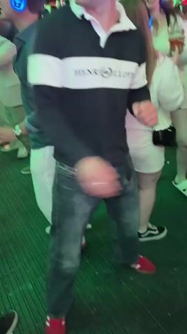 A man with brown hair wearing a dark blue and white striped polo shirt and jeans is dancing at an outdoor event. He is wearing blue headphones with glowing lights. There are other people around him also wearing headphones and dancing. The environment is lit with green lights, and there are trees and foliage visible.