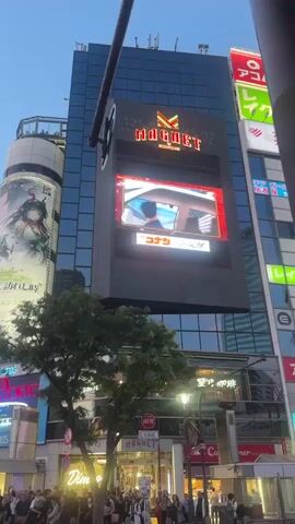 The video shows a busy street scene in Japan, with a large crowd of people waiting to cross the road. The people are diverse, with a mix of ages and ethnicities. Some of them are wearing face masks, and many are holding phones. The buildings in the background are tall and covered with advertisements, including anime characters, brands like Coca-Cola and Starbucks, and other commercial signs. The sky is light blue, indicating it's either dawn or dusk. The street is well-lit, with streetlights and the glow from the many electronic billboards. The overall atmosphere is bustling and urban.