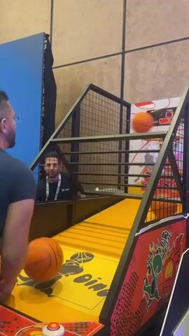 A man with glasses and a dark blue shirt is playing an arcade basketball game. He is holding an orange basketball in his right hand, preparing to shoot it towards the hoop. The game has a yellow court with red accents and graphics of cartoon characters. Another orange basketball is visible on the court near the man, and a third basketball is in the air, heading towards the hoop. A digital timer on the game screen shows 0:02. The background appears to be a wall with a textured finish. The lighting suggests an indoor environment, likely an arcade or entertainment center.