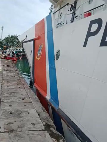 The camera angle is angled upward, showing the side of a ship docked against a concrete pier. The ship is white with a blue and red stripe running along its side. Black lettering spells out "PENG" on the side of the ship. A partially visible banner with an image of a boat labeled "KPMP" hangs from the ship. The ship is close to the pier, with minimal space between them. The pier is gray and shows signs of weathering. The background shows the sky and some greenery.