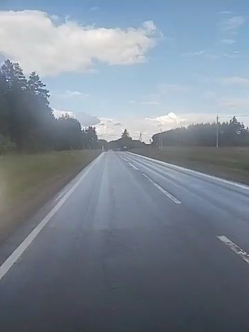 The video shows a view from a vehicle traveling on a wet asphalt road. The road is marked with white lines. On the left side of the road, there are trees and grass. On the right side, there are also trees and some utility poles. The sky is blue with scattered white clouds. The lighting suggests it is daytime. There are no other vehicles or people visible in the frame. The camera appears to be mounted at the front of the vehicle, looking forward.