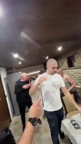 A low-angle shot captures a group of five men gathered in a room. The man closest to the camera is fair-skinned, bald, and wearing a white t-shirt and dark jeans. He holds a clear glass, drinks from it, and then gestures with the glass. To his left, a man in a black tracksuit gestures and sings into a microphone. Further back are other men, including one in a blue shirt. A table is visible to the side, with a phone and some food on it. The room has a rustic interior, with a low, dark wood ceiling, and stone walls visible in the background. The atmosphere seems lively, with music playing.