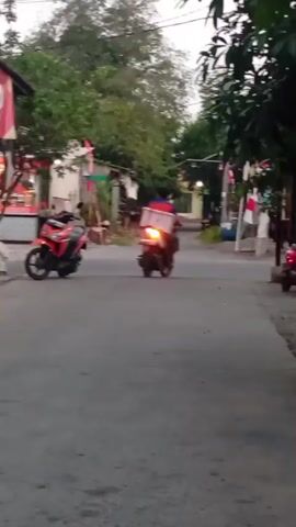 The video shows a street scene in what appears to be a residential or commercial area, possibly in Southeast Asia given the flags. Two motorcycles are visible. On the left, a red scooter is parked. Further down the street, another motorcycle, a dark color with a red seat, is moving away from the camera. The rider is wearing a dark shirt and helmet and has a box strapped to the back of the motorcycle. The street is paved, and there are trees and buildings on either side. The lighting suggests it is either early morning or late afternoon.