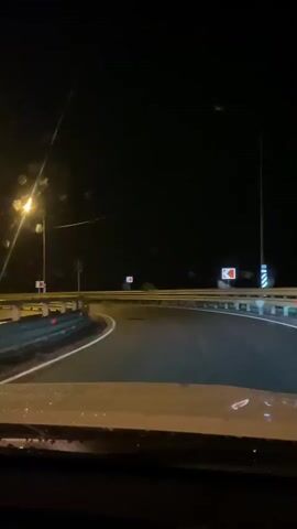 The video shows a first-person perspective from inside a vehicle driving on a dark, winding road at night. Streetlights illuminate parts of the road, casting a yellow glow. Raindrops are visible on the windshield, creating streaks and distorting the view. Traffic signs are present along the roadside, indicating turns and potential hazards. The road appears to be a highway or a well-maintained country road, with guardrails visible on the sides. The overall atmosphere is moody and slightly eerie due to the darkness and the rain.