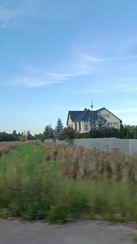 The video shows a moving shot of a rural landscape under a blue sky with some clouds. A light-yellow house with a dark-blue roof and green trim is visible behind a fence. There are several evergreen trees planted around the house. In the foreground, there is tall green grass and some dry vegetation. The camera appears to be mounted on a moving vehicle, as evidenced by the motion blur in the grass and the framing of the scene. The overall impression is of a quiet, suburban or rural setting.