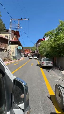 A van drives down a narrow, tree-lined street with shops on either side. Yellow arrows painted on the road indicate traffic direction. Several cars are parked along the street and a few people are walking. The sky is blue and the sun is shining.