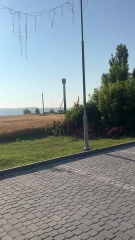 A video captured at an outdoor location on a clear day, shows a paved walkway with cobblestone pattern. A street lamp stands to the right of the frame. In the background, a field of ripe wheat stretches into the distance. Power lines and poles are visible crossing the landscape. A tall water tower is also in the background. The sky is clear and blue, with the sun shining brightly on the left side of the frame. Overhead, a string of decorative lights is hung, partially obscuring the sky. The overall scene depicts a rural or semi-rural environment under bright sunlight.