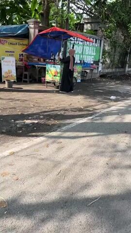 The media file depicts a street scene in Kludan, East Java. The main subject is a woman dressed in a black abaya and a hijab, standing behind a food cart. The cart is colorful, with a red and blue awning. It appears to be a vendor stall, possibly selling drinks and snacks. There are various signs on the cart, though the text is partially obscured. One sign, yellow with red lettering, mentions "HAUS ?" and "Jus Jus." Another, larger sign behind the cart, is green with white text that reads "SUBAJA FOOTBALL CULTURE."

The scene is outdoors, on a paved surface that looks like a road or pathway. The ground is covered with a mix of dirt, leaves, and some litter, suggesting it's an unkempt area. Sunlight is present, casting shadows and indicating it is daytime, likely morning or afternoon. The weather appears to be fair. Trees and foliage are visible in the background, framing the scene and suggesting a semi-urban or rural environment. The overall atmosphere seems ordinary, with no indication of any specific event or strong emotions. The focus is on the vendor and her stall, highlighting a common everyday activity of street vending in the region.