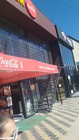 The video shows an outdoor seating area of a restaurant with a red Coca-Cola branded umbrella. In the foreground, a person's back is visible, with their hair tied in a ponytail and adorned with white star-shaped clips. Behind them, an orange soft-serve ice cream machine is positioned under the umbrella, next to a display stand showcasing images of food items like fried chicken, burgers, and fries. The restaurant facade features large glass windows and a metal staircase leading to an upper level. Several cardboard boxes are also visible on the ground. The overall setting appears to be a commercial establishment, likely a fast-food or casual dining restaurant, on a sunny day.