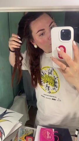 A woman stands in front of a mirror taking a selfie with her white iPhone. She has long, reddish-brown hair, with some strands styled into tight curls. She is wearing a white T-shirt with a yellow and black graphic print, and pearl stud earrings. Her fingernails are painted with a white and gold design. She is touching her hair with one hand as she takes the selfie. The background is a cluttered space, possibly a dressing room or small office. There is a sheet of paper with a colorful drawing on a desk below the mirror. Other items visible on the desk include a small pink container with a handle, a book, and a makeup product. The wall behind the mirror is painted in a faded greenish-blue color. The mirror reflects the woman and some objects in the room, adding to the cluttered appearance.
