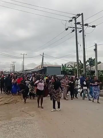 A group of people are walking and dancing down a street. The people are dressed in a variety of clothing, many wearing traditional African attire. There are buildings and power lines visible in the background. The sky is overcast.