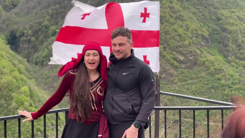 A medium shot shows two people standing at a viewpoint, a woman in traditional Georgian clothing and a man in a black Nike jacket. Behind them waves a Georgian flag. A mountainous, green landscape forms the backdrop. The woman in the traditional outfit speaks while holding the man's hand, seemingly addressing a group of people who are partially visible at the edges of the frame. The man looks towards the group, listening. The overall atmosphere suggests a cultural presentation or tour.