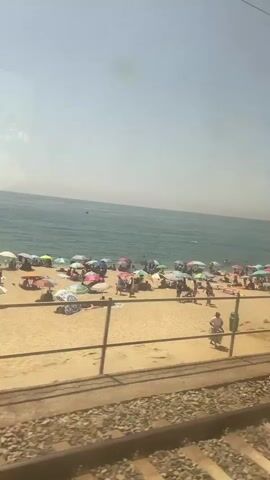 The video shows a beach scene from the perspective of a moving train. The train tracks and ballast are in the foreground. Beyond the tracks, there is a sandy beach with numerous people and colorful beach umbrellas. People are seen sunbathing, walking, and swimming in the turquoise water. The sea stretches to the horizon under a clear sky. The train moves from left to right, gradually revealing more of the beach and the trees lining the shore.