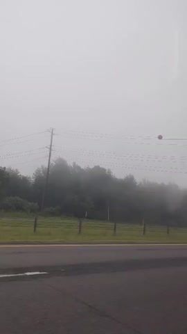 This media file captures a view from a vehicle driving on a road in a foggy environment. The scene is dominated by a thick fog, obscuring much of the background and creating a hazy atmosphere. In the foreground, a paved road with yellow and white lane markings is visible. Beyond the road, there's a grassy area bordered by a fence, and further back, a dense line of trees is mostly hidden by the fog.

Several man-made objects are present: power lines stretch across the frame, supported by utility poles. A red spherical object is suspended from one of the power lines. A dark-colored vehicle is partially visible in the left side of the frame, and a white SUV is seen in the distance on the road.

Further along the road, a green road sign indicating "EXIT 3" with an arrow pointing to the right is clearly visible. Below this sign are two yellow diamond-shaped warning signs. Another green road sign is faintly visible in the distance on the left.

The weather is clearly foggy, significantly reducing visibility. The time of day is difficult to determine precisely due to the fog and overcast conditions, but it appears to be daytime. The overall mood is somewhat muted and mysterious due to the fog. The location is identified as Tobyhanna Township, United States of America. There are no people directly visible in the frame, but reflections on the vehicle's window suggest the presence of someone inside.