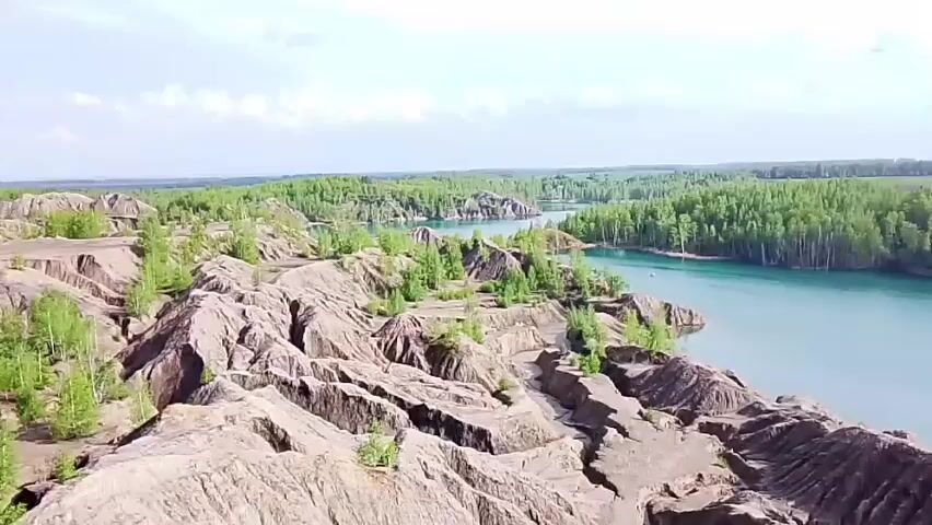 The video displays an aerial view of a landscape featuring a body of water with a striking turquoise hue. The water is surrounded by a combination of land formations, including undulating, brown, dirt hills that appear to be relatively barren, punctuated by small patches of green vegetation. Across the water, a dense forest of green trees extends into the distance, merging with a flat, grassy plain under a sky lightly brushed with clouds. The scene suggests a contrast between the rugged terrain and the lush greenery, all under a bright, overcast sky.