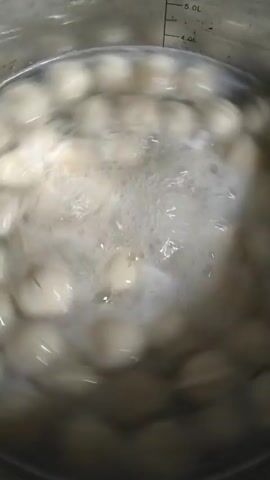 A close-up shot shows many small, round, pale objects, likely dumplings or tortellini, submerged in boiling water within a metal pot. The water is actively bubbling and frothing, indicating a high temperature. The movement of the water causes the objects to shift and tumble. A measurement line is visible on the upper right interior of the pot, marked at "4.0L". The lighting is overhead, creating a bright, slightly diffused illumination of the scene.