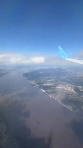 The video depicts an aerial view from an airplane window. The sky is blue with some scattered white clouds. A portion of the plane's wing, painted white with a light blue tip, is visible in the upper right corner. Below, a wide river stretches across the landscape. A bridge spans the river, connecting land on either side. The land consists of a mix of green areas, likely vegetation, and what appear to be urban or suburban developments with roads. The river water appears murky. The overall scene is calm and serene.