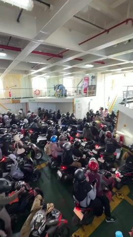 The video shows the interior of a ferry, packed with numerous motorcycles and their riders. The motorcycles are arranged in rows, filling most of the available space. People, mostly wearing helmets and casual clothing, are seated on their motorcycles, facing forward and waiting. Some are looking at their phones, while others appear to be talking to each other. The environment is industrial, with visible structural beams, pipes, and fluorescent lighting on the ceiling. Yellow markings are visible on the floor, indicating parking lanes. The overall impression is one of a busy, crowded ferry preparing for or in the midst of a journey.