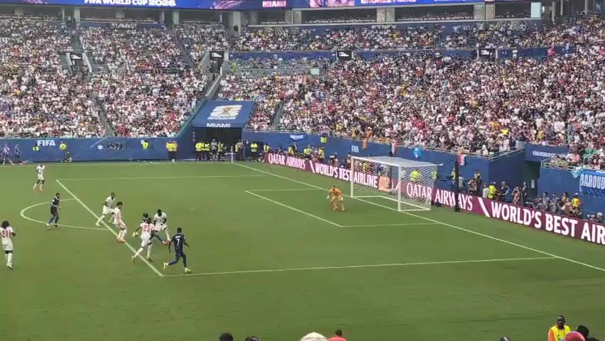 A wide shot captures a soccer match in a packed stadium. The field is green, with white lines marking the boundaries and penalty areas. Players in white and dark blue uniforms are visible on the field, actively engaged in the game. A goalkeeper in a yellow jersey stands in front of a white-netted goal. The stands surrounding the field are filled with a large crowd of spectators, many of whom are holding up their hands. Banners and advertisements are displayed around the stadium, including one that reads "QATAR AIRWAYS WORLD'S BEST AIRLINE." The scene appears to be set during daylight hours.