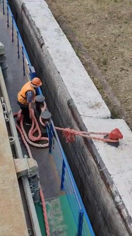 From an elevated angle, the video shows a person in a hardhat and orange vest securing a thick, red rope around a series of bollards on a boat. The person is on the port side of the boat, working to control the rope, which extends from the boat to a red bollard on the cement bank next to the boat. The boat is in a narrow channel with cement walls on both sides, separating it from the surrounding grassy area. A blue railing separates the person and the working area from the open edge of the boat.