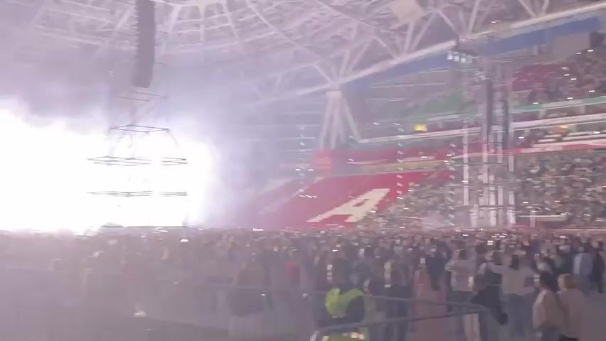The video shows a crowded stadium with people holding up their phones, illuminating the stands. Green lights and smoke are visible, creating a dramatic atmosphere. A large red banner with white lettering is unfurled across one section of the seating. The audience appears to be excited and engaged, with many people standing and cheering. The stadium architecture suggests a large sporting or entertainment venue.