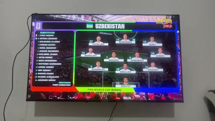 This media file shows a television screen displaying information about a soccer match featuring Uzbekistan. The screen is divided into sections. On the left, a list of substitute players for Uzbekistan is shown, along with the head coach, Fabio Cannavaro. The central and right portions of the screen display the starting lineup of the Uzbekistan team, with player names and numbers overlaid on images of the players. The background graphics suggest a digital representation of a soccer field. The text "UZBEKISTAN" is prominently displayed at the top, along with the FIFA logo and "LIVE." Below the team lineup, the text "FIFA WORLD CUP 2026™" is visible. The second part of the media file shows a wide shot of a packed soccer stadium filled with spectators wearing red. The field is green, and the stands are full, indicating a live sporting event. The overall atmosphere appears to be energetic, typical of a major sporting event. No specific time of day or weather conditions are evident, but the setting is clearly a professional soccer stadium. While the text on the screen indicates "FIFA WORLD CUP 2026™," the context of Mashrab, Uzbekistan, is not directly verifiable from the visual information alone.
