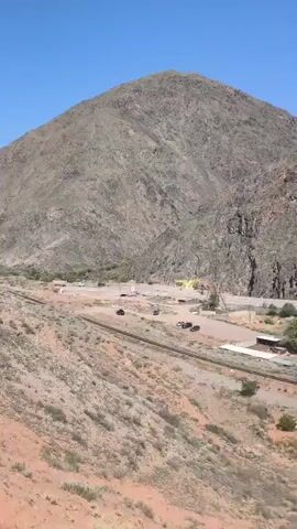 The video presents a static, wide-angle view of a mountainous landscape on a bright, clear day. In the foreground, the terrain is characterized by reddish-brown, arid soil dotted with sparse vegetation. A single set of railroad tracks runs horizontally across the midground, with several buildings and trees clustered along the tracks. A road and a few cars are seen further back. In the background, a large, rocky mountain dominates the scene, its surface textured with shades of gray and brown. The sky is a clear, unblemished blue, suggesting a dry and sunny climate.