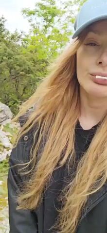 Here's a concise description of the video:

A woman with long, reddish-blonde hair is walking along a path, wearing a light blue baseball cap and a black jacket. She has a thin, white object, possibly a toothpick or a candy stick, held in her mouth. The path appears to be gravel or dirt and winds alongside a hillside covered in green trees and vegetation. The sky is overcast. The camera seems to be held by the woman herself, giving a first-person perspective. The focus is mainly on her face as she walks.