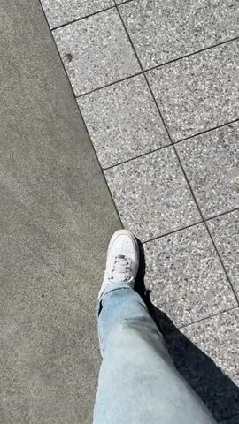The video begins with a first-person perspective shot of a person's legs and feet wearing white sneakers and blue jeans walking on a textured grey tiled surface. A long shadow is cast to the right. The camera then pans up and to the right, showing a set of concrete stairs with metal railings. A person wearing a black shirt and blue jeans walks up the stairs away from the camera. The camera continues to pan, revealing an outdoor seating area of a coffee shop. Two women are seated at a table under a white umbrella. One woman has blonde hair and wears a white shirt and blue jeans, while the other has brown hair and wears a black dress. A third person, the man in the black shirt, walks past the seating area towards the entrance of the coffee shop, which has the words "KIT COFFEE" visible on the glass door. The scene is outdoors on a sunny day.