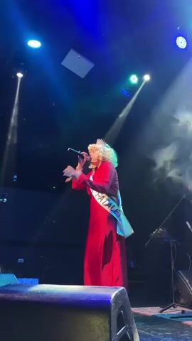 A person with blonde hair, wearing a crown and a red dress, stands on a stage singing into a microphone. A sash with the words "MAMA FATALE" is draped across their chest. The stage is lit with blue and white spotlights, and there is smoke or fog on the left side of the stage. The person is holding the microphone with their right hand and gesturing with their left. They appear to be performing for an audience, though the audience is not visible in the frame. There are speakers and other stage equipment visible in the background.