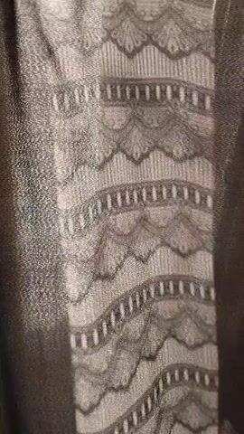 This is a close-up, vertical shot of a black fabric with a lace panel. The lace has a wavy pattern with vertical lines and decorative edges. The fabric to the left of the lace appears to be slightly textured and shimmery. The fabric to the right is a plain black material. The shot is static and the lighting is dim, suggesting it may be indoors.