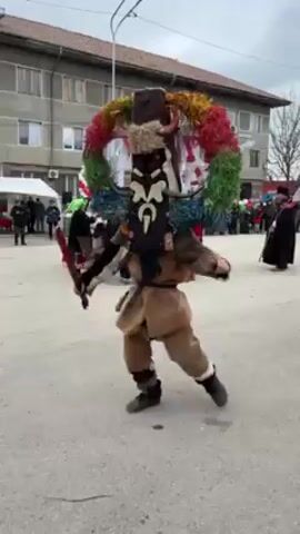 People are dressed in traditional Bulgarian costumes and masks. They are walking, dancing, and socializing in a town square. Buildings and street lights are in the background. It is daytime.