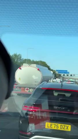 The video shows a view from inside a car stuck in traffic on a sunny day. The foreground is dominated by the rear of a black car with a license plate reading "LE75 DZX". The car's brake lights are illuminated. Behind this car, a large white tanker truck is visible, carrying what appears to be a liquid or gas. The truck has hazard symbols on its rear.

The scene is set on a multi-lane highway with other vehicles also stopped in traffic. Overhead signs are visible in the distance, suggesting a major road. Trees line the side of the highway. The sky is clear blue, indicating good weather and likely daytime. The overall mood is one of frustration or patience due to the traffic congestion. No people are clearly visible, but the presence of multiple vehicles implies drivers and possibly passengers.

Some text is visible on the rear window of the black car. It appears to be stickers, with some words in English that are partially obscured and reversed due to being seen through the glass. One sticker appears to be a red square with white text.

The location context is Three Rivers, United Kingdom, which is a district in Hertfordshire, England, known for its urban areas and proximity to London, suggesting this could be a common commuter route or a major artery.