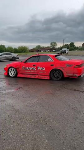 A bright red Toyota sedan with "Ceramic Pro" displayed on its side is drifting on a wet, muddy surface. The car leaves dark tire tracks on the ground as it moves. Another silver sedan is parked to the side. A person dressed in a white shirt and hat, and carrying a tennis racket, stands by the silver sedan. The sky is overcast, and trees are visible in the background.