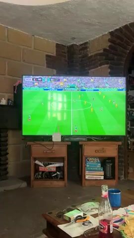 The video displays a large television screen showing a soccer match. The score is tied at 0-0. The game is between Mexico (MEX) and South Africa (RSA). The time on the clock is 2 minutes and 25 seconds. A player in a green and white jersey with the number 3 on his back is visible on the field, dribbling the ball. The stadium is filled with spectators wearing green, suggesting a strong Mexican fan presence. The TV is positioned on top of two wooden nightstands. The room appears to be a living area with brick walls and a stone fireplace. In the foreground, on a coffee table, are various items including a bottle of red liquid, a blue mug, sunglasses, a pack of cigarettes, and what appears to be a lighter. The lighting in the room suggests it is evening or indoors with artificial light.