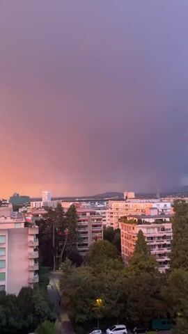This is a vertical, aerial shot overlooking a city during a sunset. The sky is a dramatic gradient of purple, pink, and orange, with dark storm clouds on the right side of the frame. Below the sky, the tops of several multi-story buildings are visible, some with balconies and others with flat roofs. A line of trees, mostly dark green, partially obscures the lower levels of the buildings. In the distance, a hazy mountain range is visible under the colorful sky. There are no people or discernible actions captured in this video.