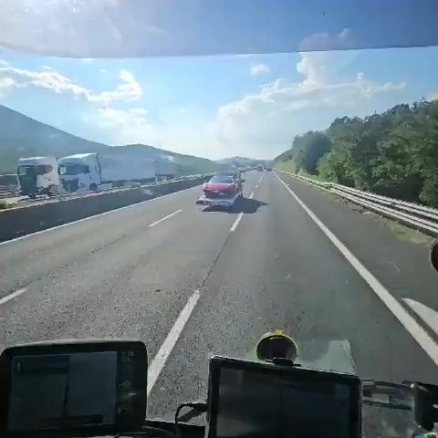 The video shows a red sports car being transported on the back of a tow truck on a highway. The car appears to be a Ferrari. The tow truck is driving on the right side of the road, in the middle lane. There are other vehicles on the highway, including trucks and cars. The weather is sunny, and the sky is blue with some clouds. The road is flanked by hills and trees. The video is filmed from the perspective of another vehicle, presumably following the tow truck.