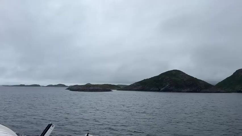 The video shows a view from a boat moving through water towards a cluster of green, hilly islands under a cloudy, overcast sky. The water is dark and choppy. The boat is moving away from the islands, as indicated by the wake visible behind it. The landscape is rugged and sparsely vegetated. The sky is uniformly gray and appears to be covered by clouds. The video is a continuous shot without any visible people or specific objects on the boat apart from what appears to be the edge of the boat in the lower left corner. The camera seems to be positioned at a medium height relative to the water. The overall impression is one of a desolate, natural environment.