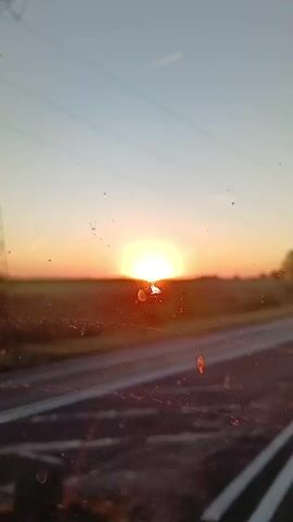 The video shows a sunset viewed from inside a vehicle. The sun is low on the horizon, casting a warm orange glow across the sky and landscape. A road with white lines is visible in the foreground, and a field of tall grass or crops stretches into the distance. Power lines and a distant transmission tower are also present in the frame. The sky is clear with a few faint contrails from airplanes. The overall impression is one of a peaceful, rural setting at dusk.